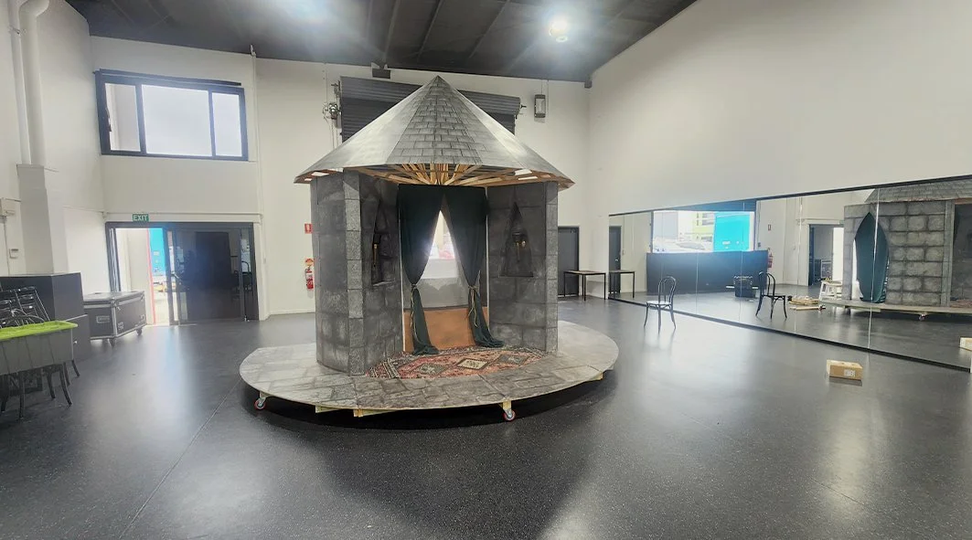Indoor scene featuring a small castle-like structure with stone walls and a conical roof, set on a circular platform with wheels, inside a spacious room with black flooring, high white walls, and several chairs and tables.