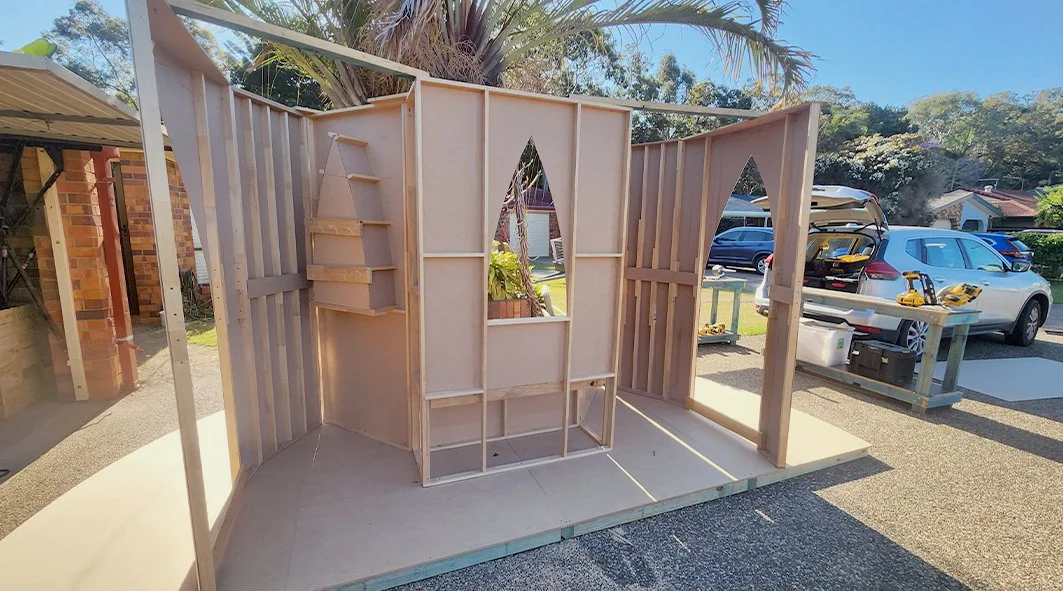 Wooden framing for a small outdoor structure under construction with an arch-shaped cutout and shelving.