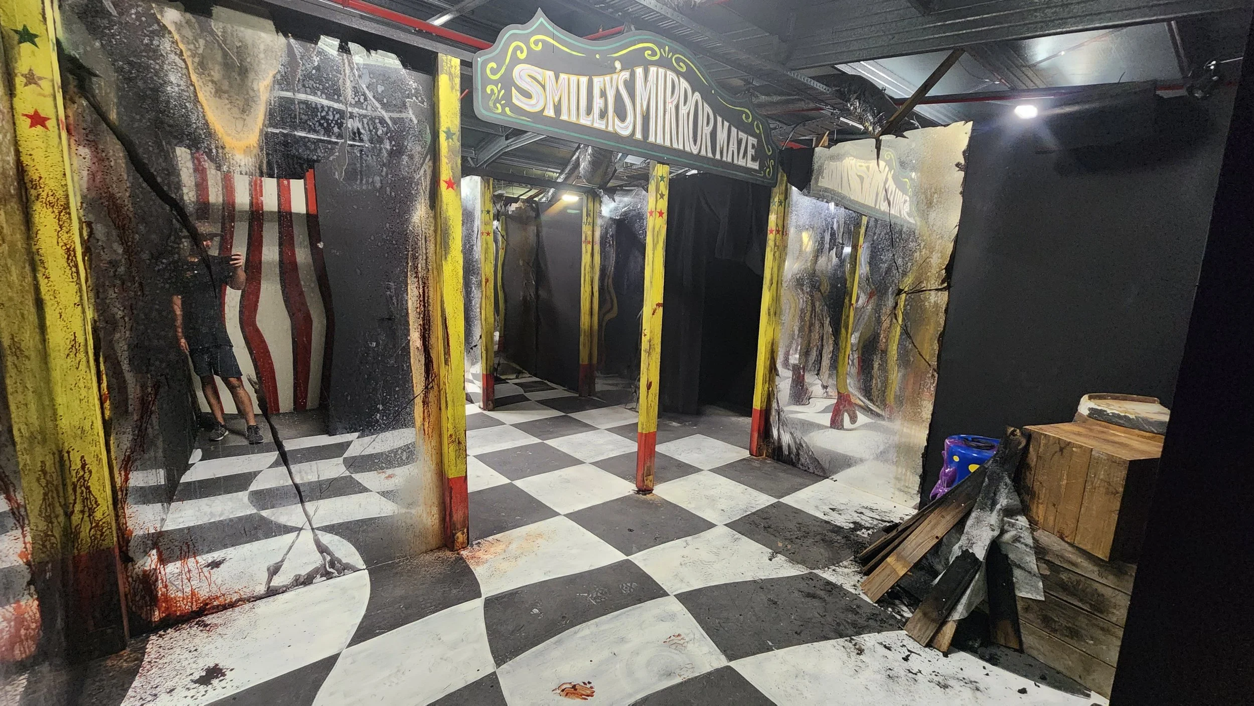 The image shows an exit area of a haunted house attraction called 'Smiley's Mirror Maze.' The area is decorated with yellow and red metal posts, some of which have been splattered with red paint resembling blood. There are black walls and a checkered floor, with the walls and posts covered in murals and designs that give a creepy vibe. A sign above reads 'Smiley's Mirror Maze,' and there are black curtains hanging at the entrance. There is also some wood and other debris on the right side of the area.