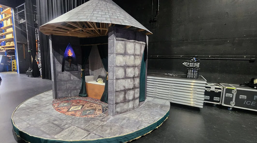 A small theater stage set with a medieval castle theme, featuring a stone wall facade, a round carpet, curtained entrance, and a throne inside, with stage equipment and storage visible around.