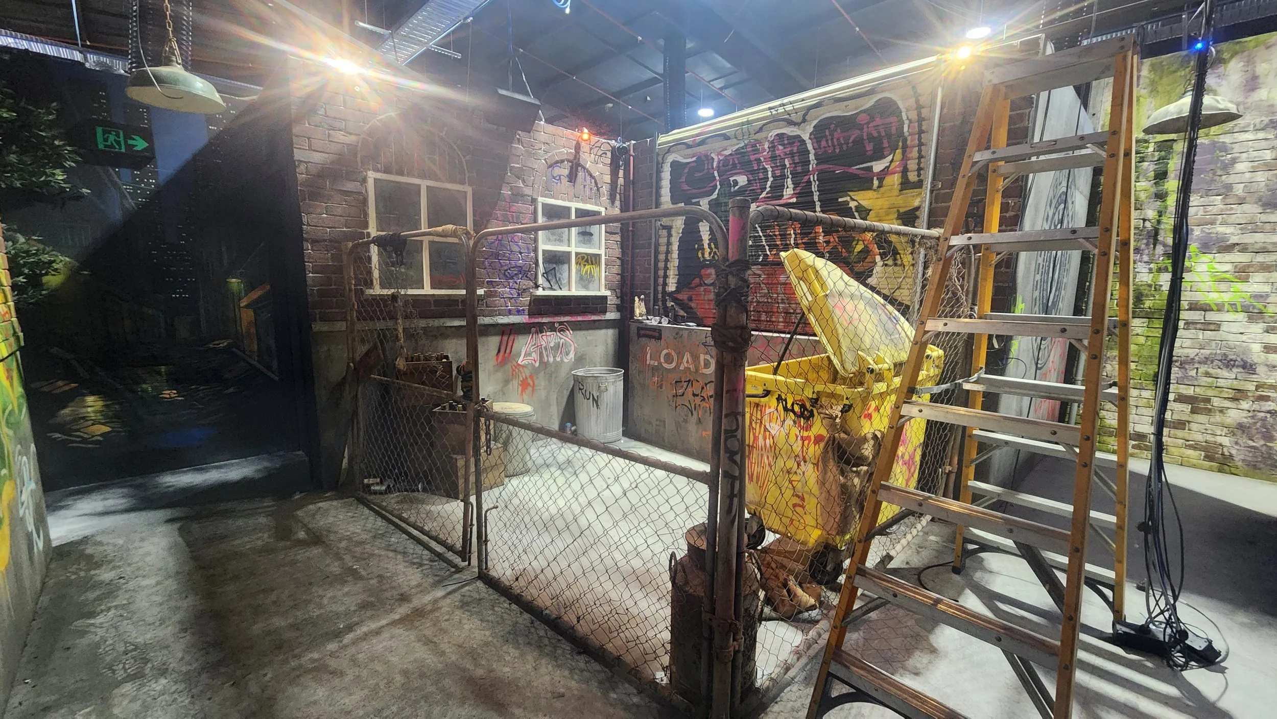 Indoor space with brick walls and graffiti art, metal fencing, a ladder, and construction equipment, including a yellow wheelbarrow and trash bin.