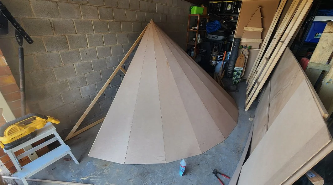 Woodworking workshop with a conical wooden structure under construction, surrounding materials, tools, and storage shelves.