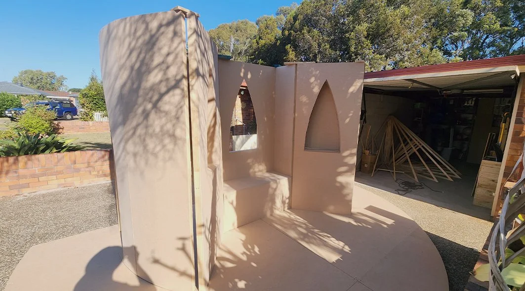 A partially constructed small structure with arched windows, made of beige material, outdoors in a backyard with a brick wall, trees, and a garage with woodworking frames and tools.