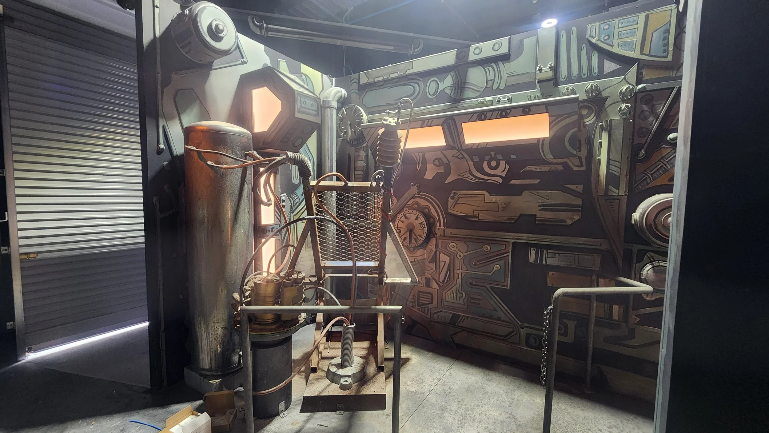 A setup with various scientific equipment including a rusted metallic tank, hoses, gauges, and a cart holding smaller devices, with a background mural featuring circuitry and mechanical designs.