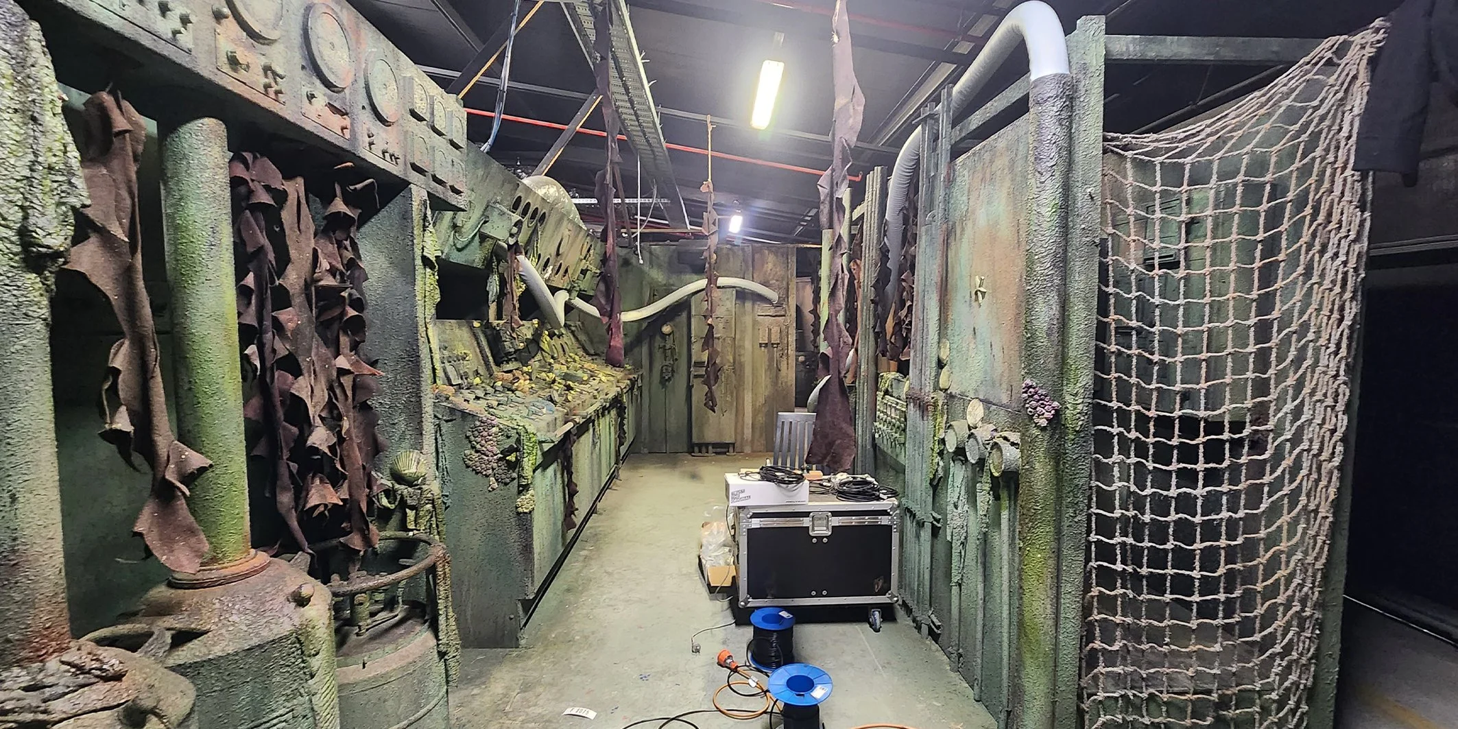 A room that looks like a set from a post-apocalyptic movie or a game, with walls covered in moss and dirt, and various objects such as pipes, wires, and machinery. There's a black box, some cables, and equipment on the floor.