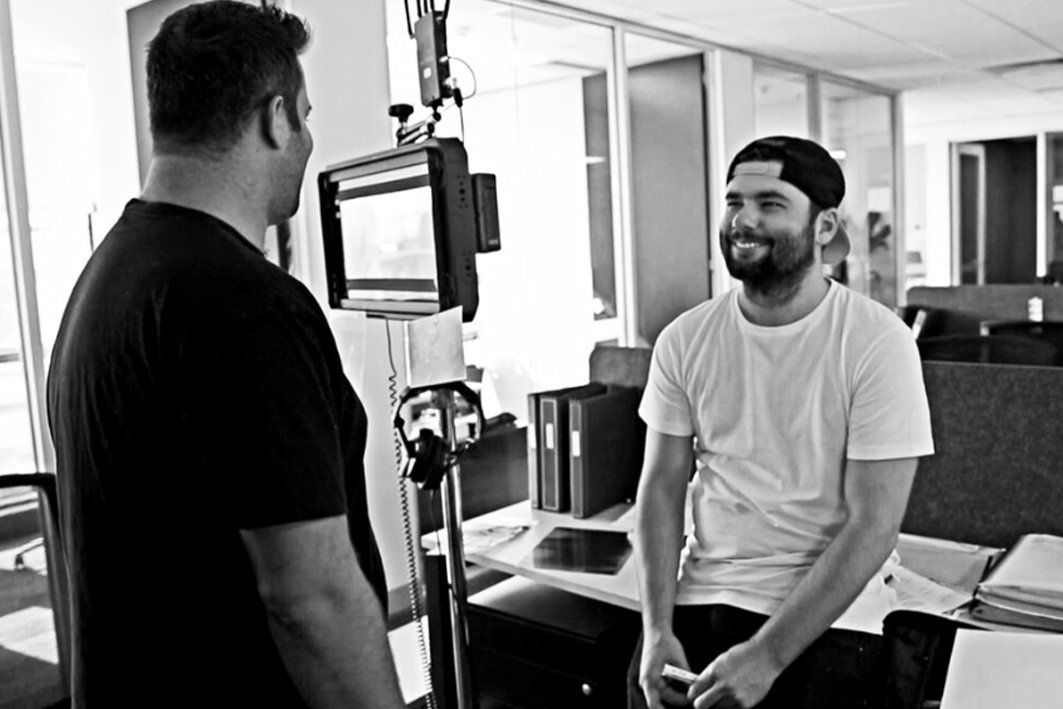 Two men in an office setting, one filming the other who is smiling and sitting on a desk.