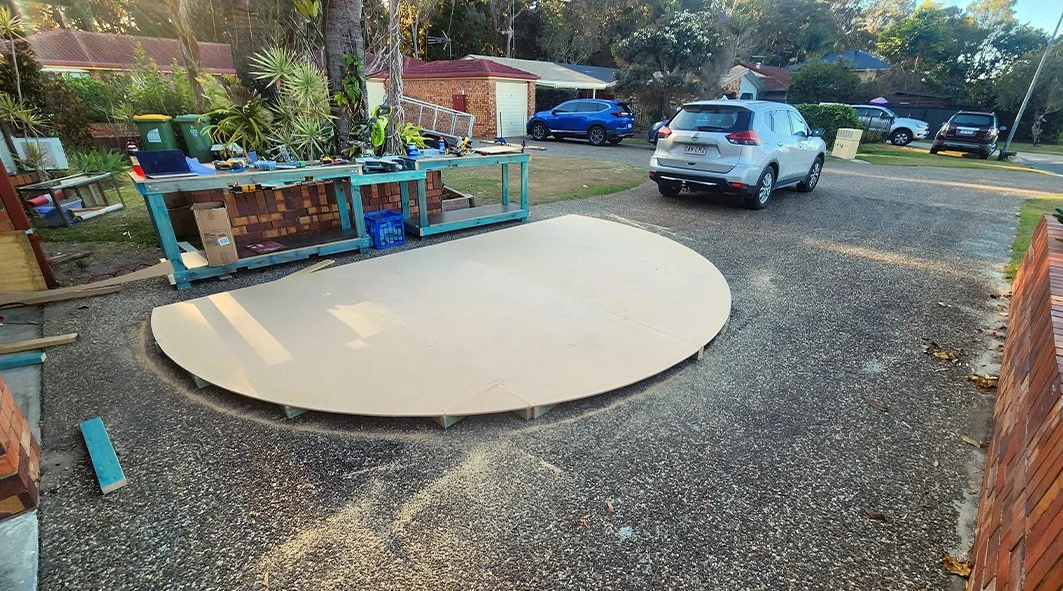 A round skateboard deck on a paved driveway, with construction tools and materials around it, next to a workbench and parked cars in a suburban area.