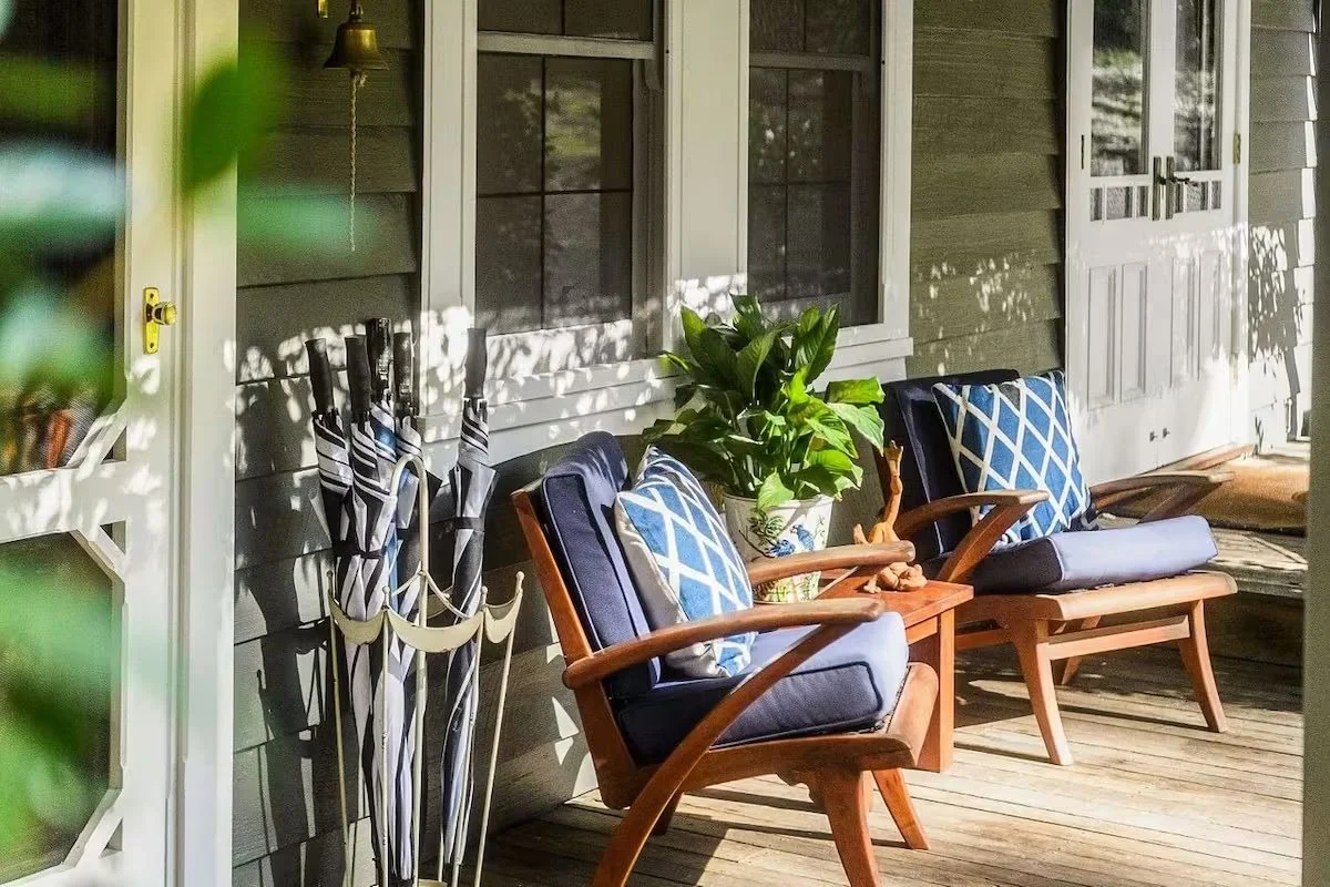Sunlit porch with two wooden chairs with blue cushions and patterned pillows, a potted plant between the chairs, and closed umbrellas stored beside the chairs.