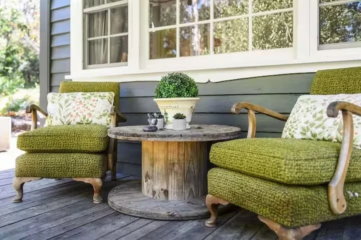 Porch with two green upholstered armchairs flanking a round wooden table holding a potted plant and decorative items, in front of a house with large windows and blue siding.