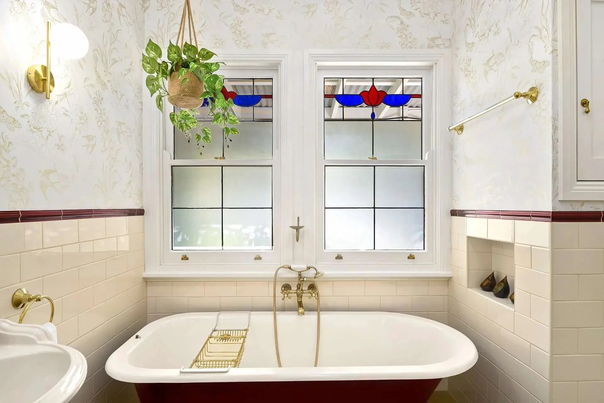 Bathroom with white tile walls, a vintage-style bathtub with brass fixtures, two stained glass windows, a hanging plant, and brass wall sconce lights.