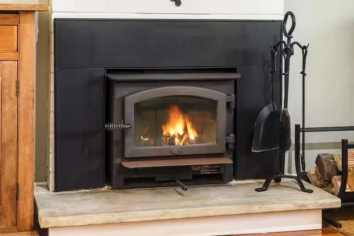 A fireplace with a fire burning inside a glass door, surrounded by a black mantel, with fireplace tools and wood logs nearby.