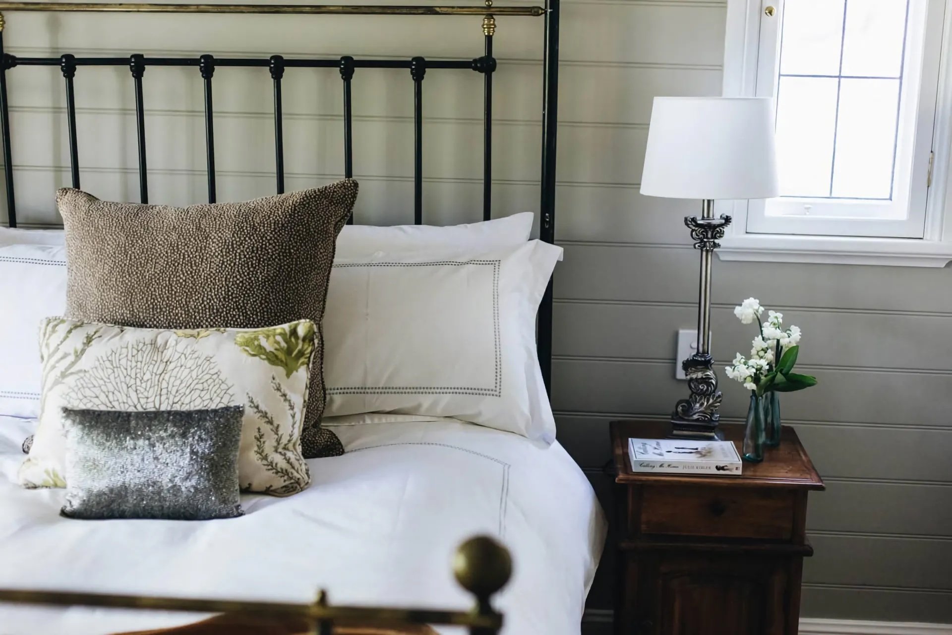 Close-up of a neatly made bed with multiple decorative pillows, including one with a textured pattern, situated next to a side table with a lamp, flower vase with white flowers, and a magazine, in a bedroom with white walls and a window letting in na