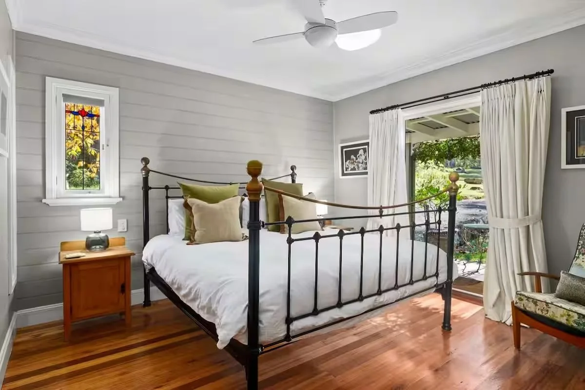 Bright bedroom with gray walls, black metal bed with white bedding and multiple pillows, wooden nightstand with a lamp and clock, hardwood floors, window with stained glass, glass door with white curtains leading to outdoor patio.