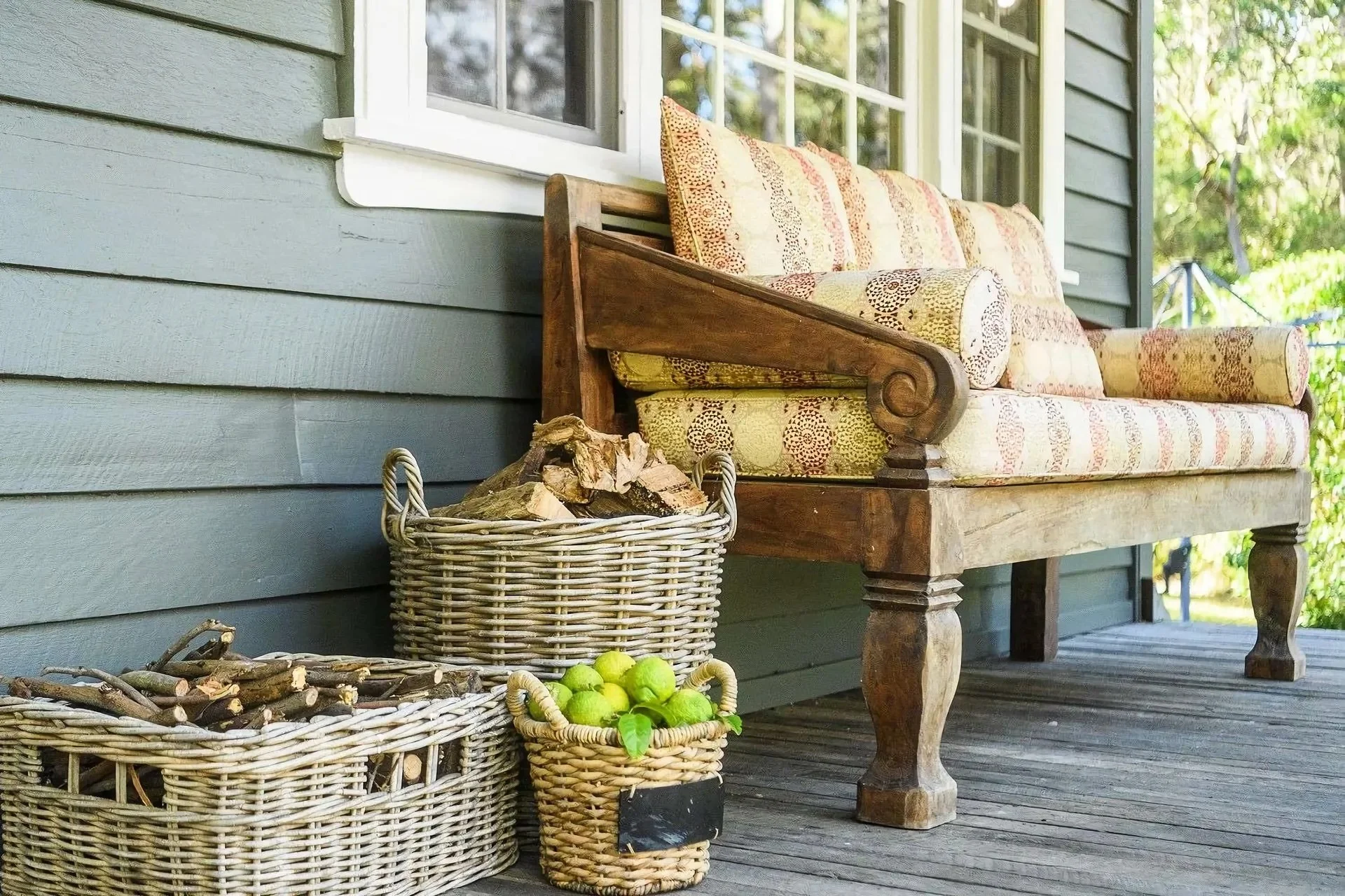 A front porch with gray wooden siding, a large window, and a wooden bench with colorful patterned cushions. Two wicker baskets contain firewood and green apples. A small blackboard sign leans against the smaller basket. The porch has a wooden floor a