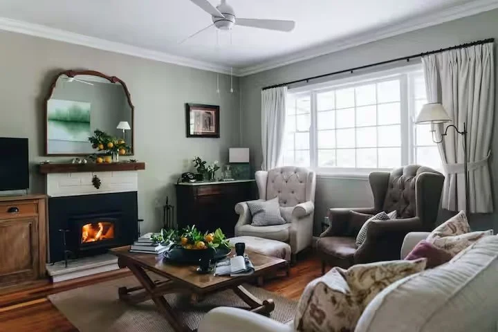 Bright living room with large window, cream-colored armchairs, and a fireplace with a mirror above it.
