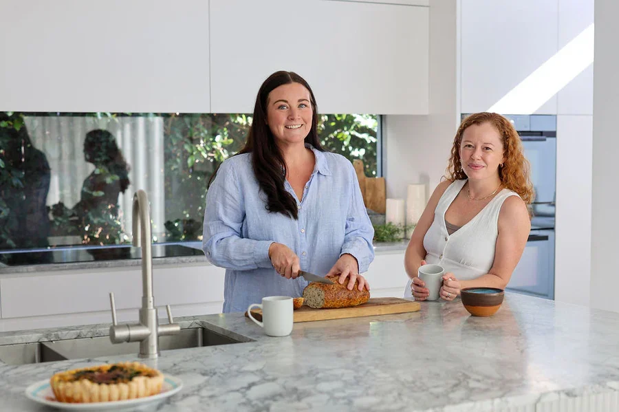 Two women in a modern kitchen, one slicing bread and the other holding a mug, with a pie on the countertop.