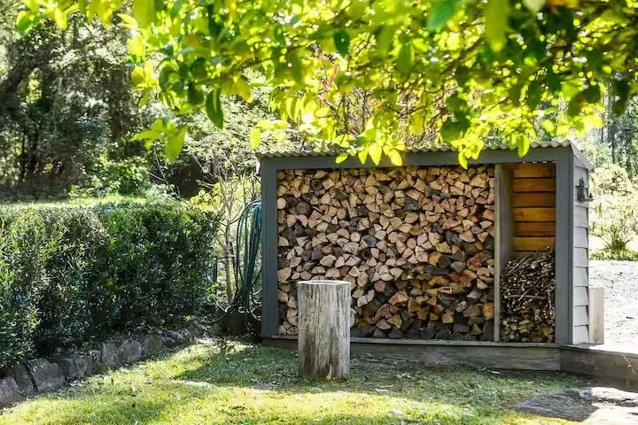 A wood storage shed filled with chopped firewood, situated in a yard with grass, a tree, and shrubs.