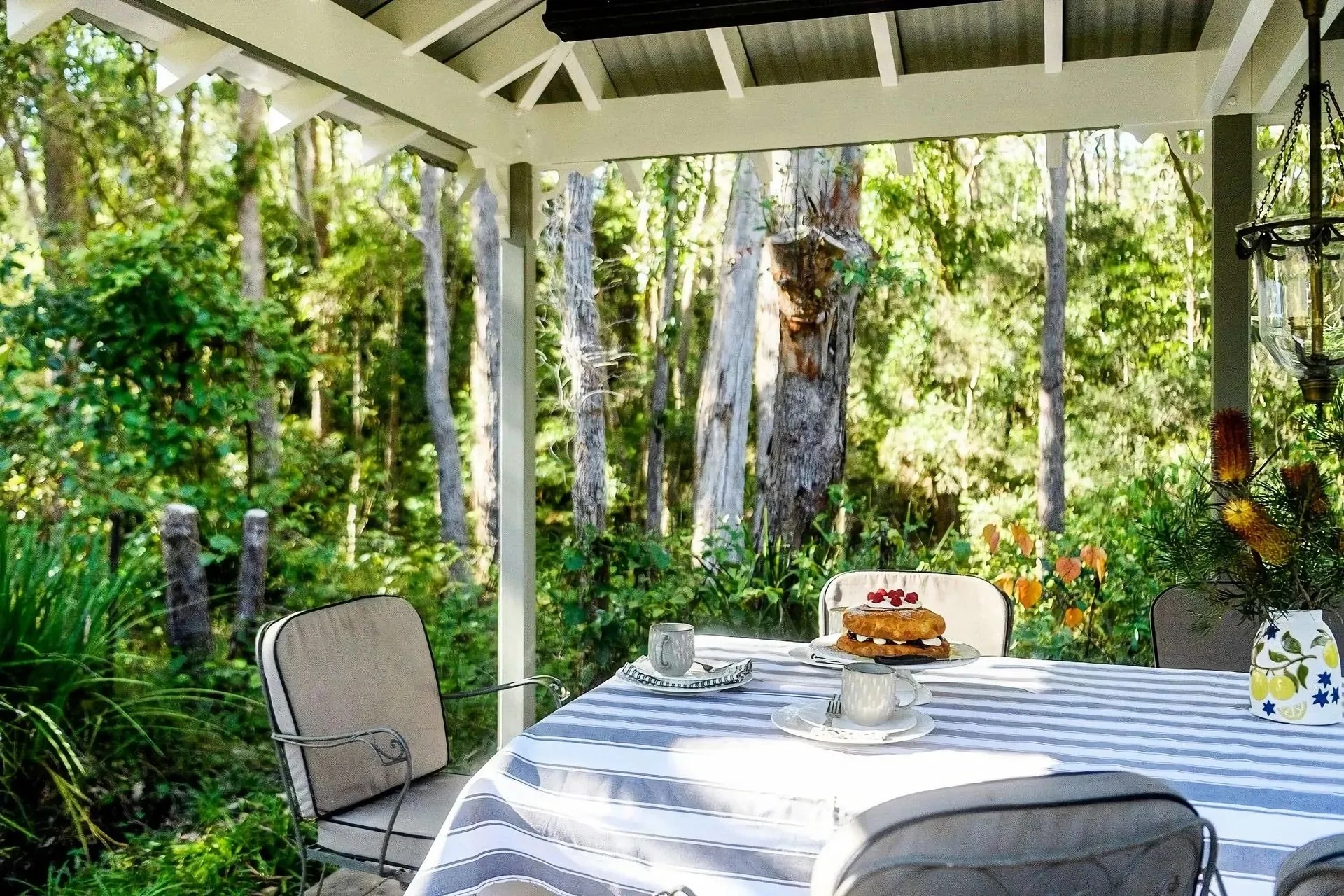 Outdoor dining table with a blue and white striped tablecloth, set with plates, cups, and a sandwich, on a porch with chairs, overlooking a lush, wooded backyard with trees and greenery.