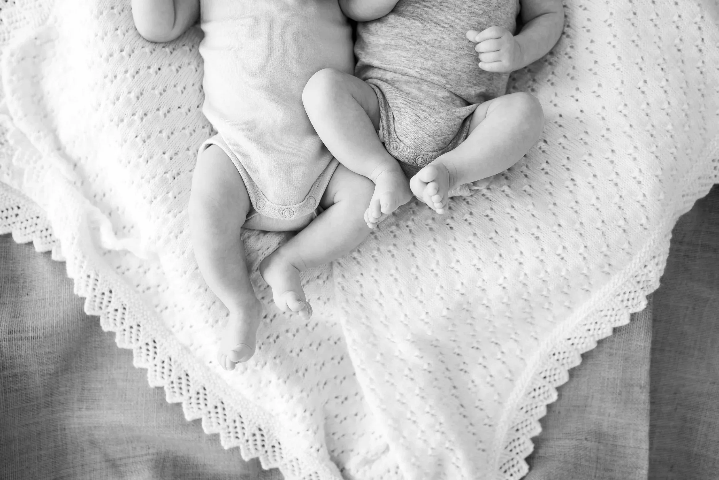 Black and white photo of two babies lying on a crocheted blanket, touching each other.