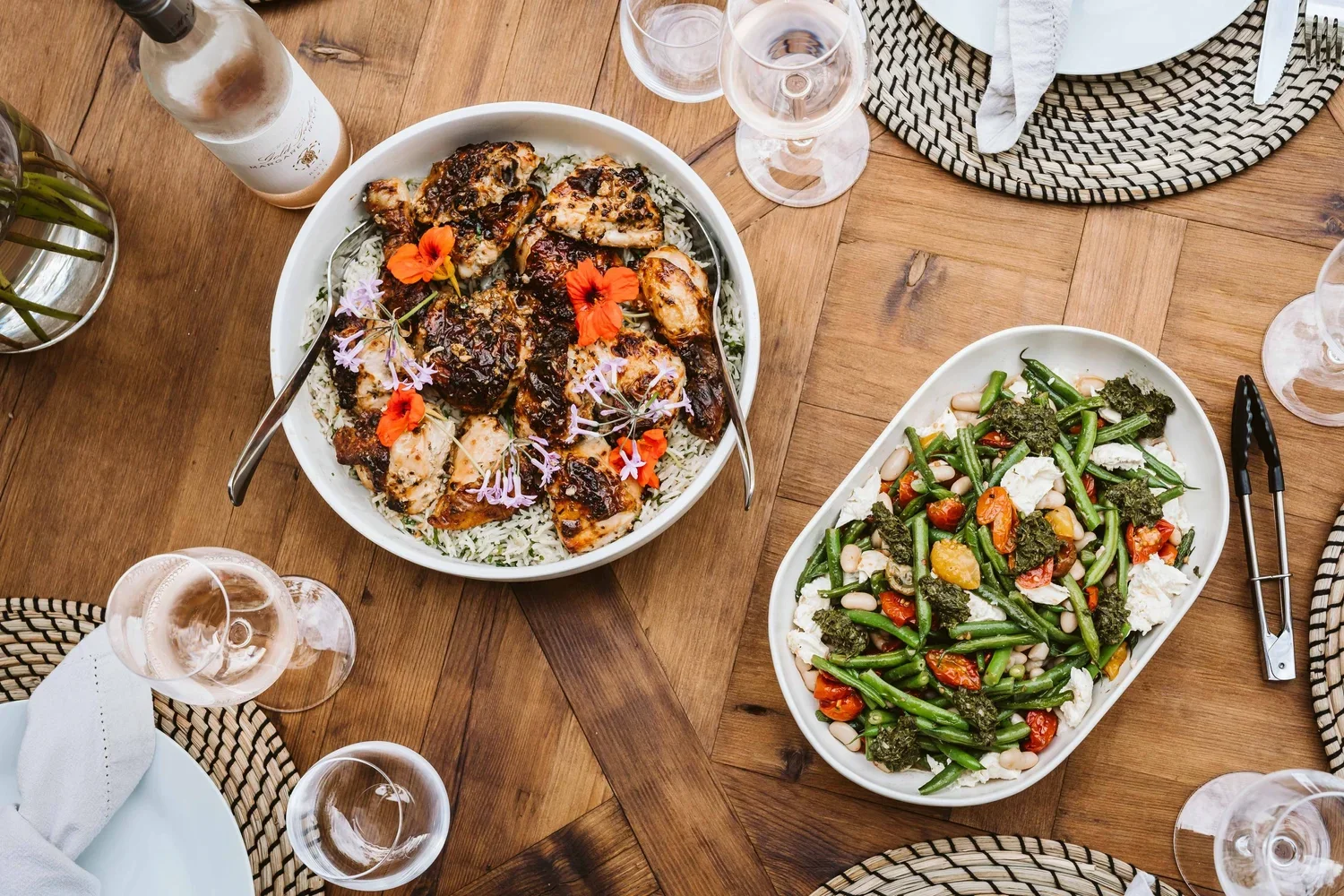 Two large serving dishes on a wooden dining table, one with grilled chicken topped with herbs and edible flowers over rice, the other with green beans, cherry tomatoes, white beans, and leafy greens, surrounded by wine glasses, plates, and cloth napkins.
