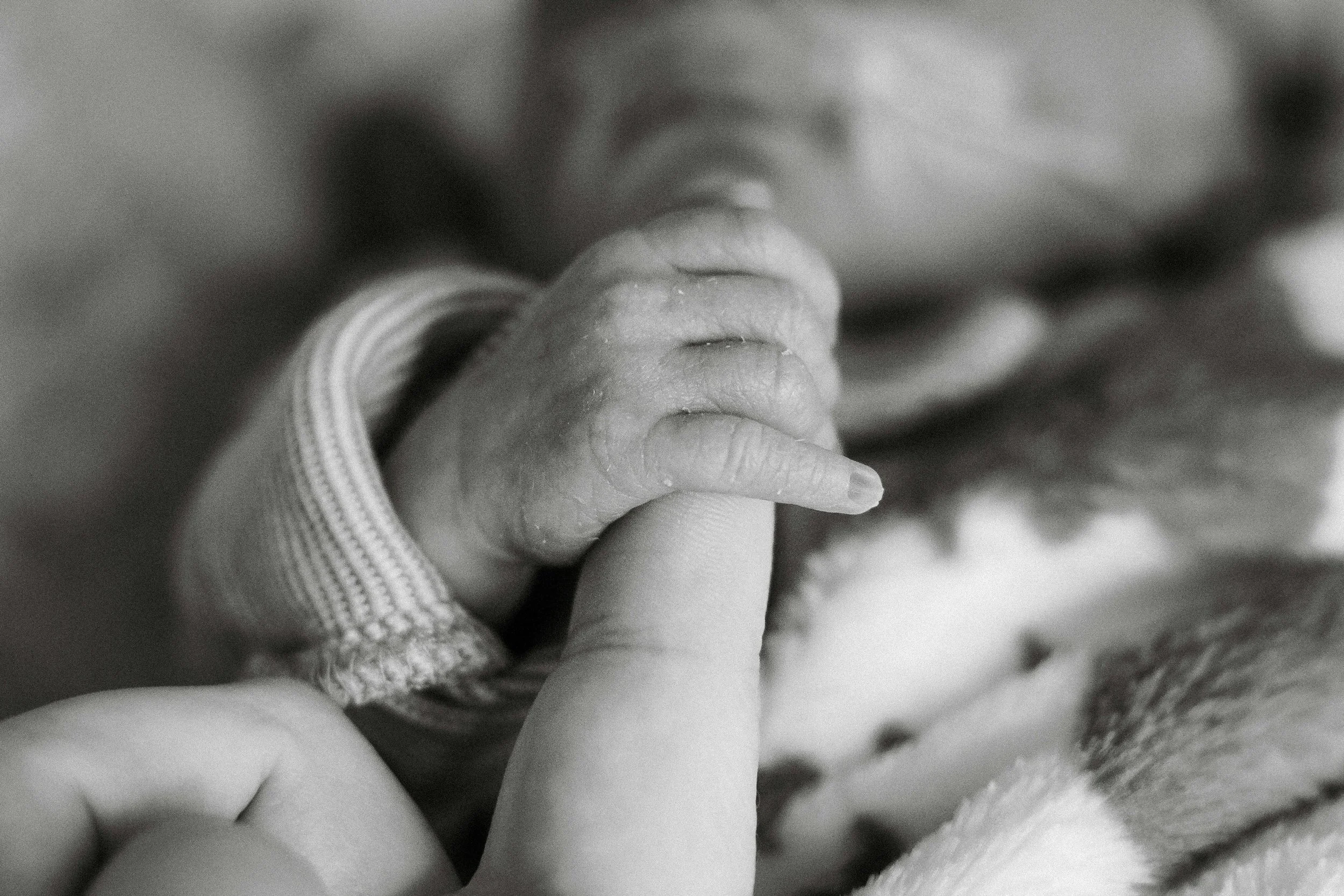 Close-up of a baby's hand gripping an adult's finger, notable for its softness and tiny size, in black and white.