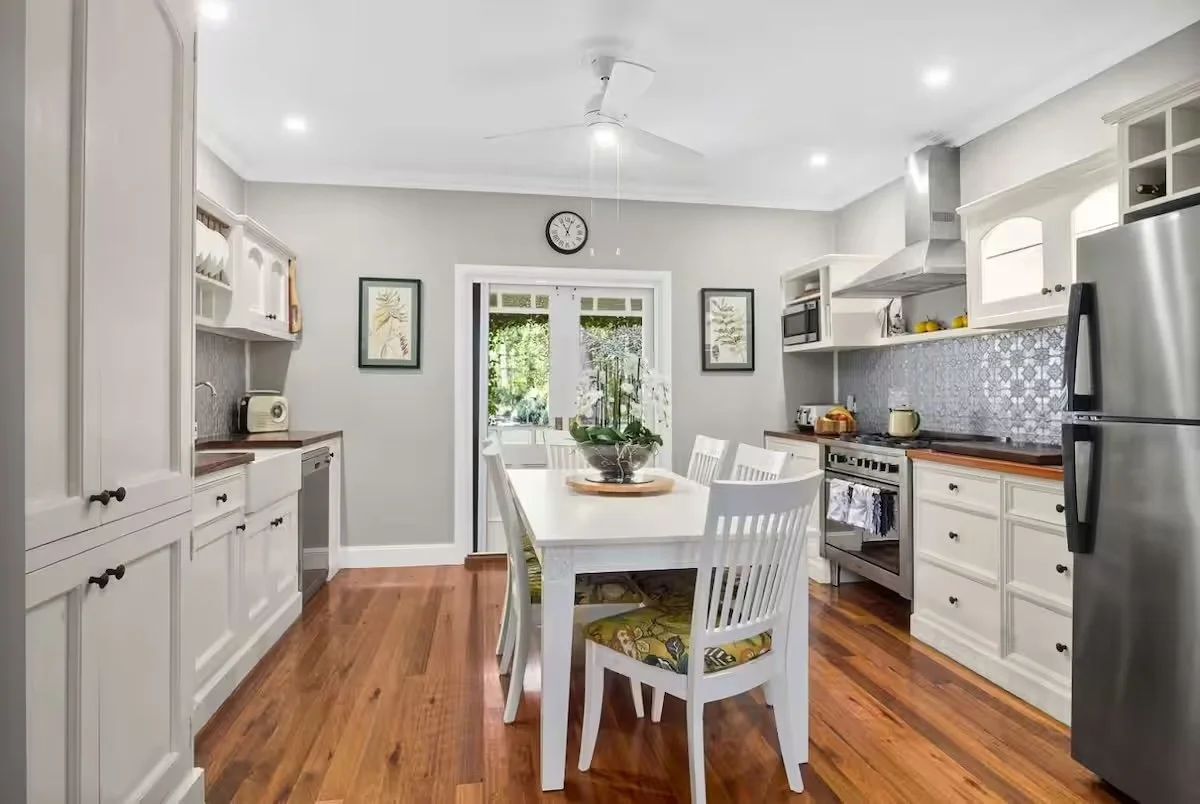 Kitchen with white cabinets, a dining table with six white chairs, stainless steel refrigerator, wood flooring, and a window with greenery outside.
