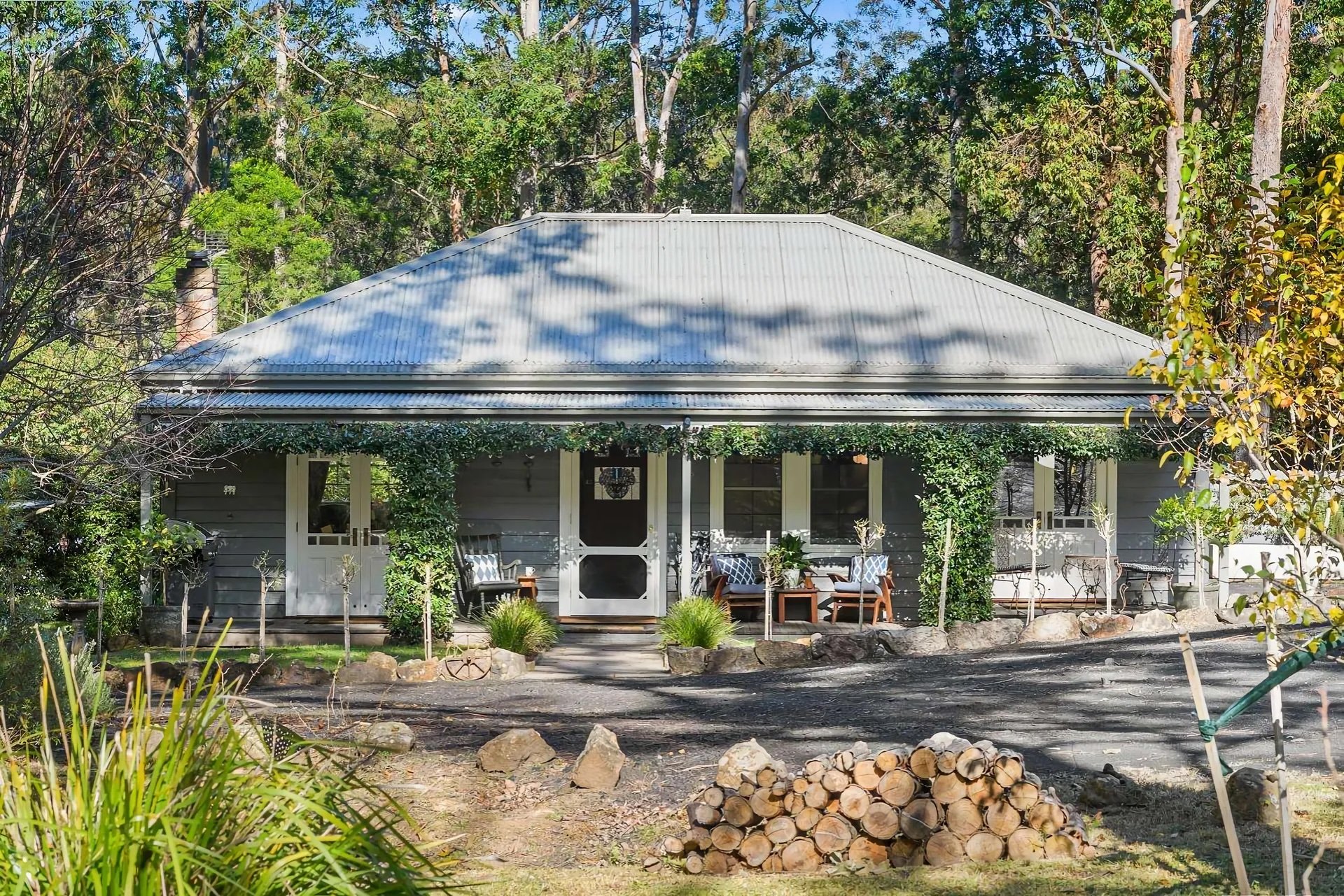 Front view of a house with a gray roof, tropical plants, trees, and a small seating area in the garden.
