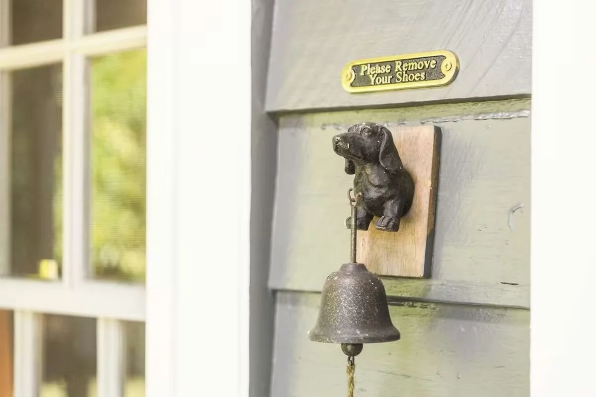 A decorative doorbell with a dog figure holding a bell on a wooden base, mounted on a green-painted door with a brass sign above that reads 'Please Remove Your Shoes.'