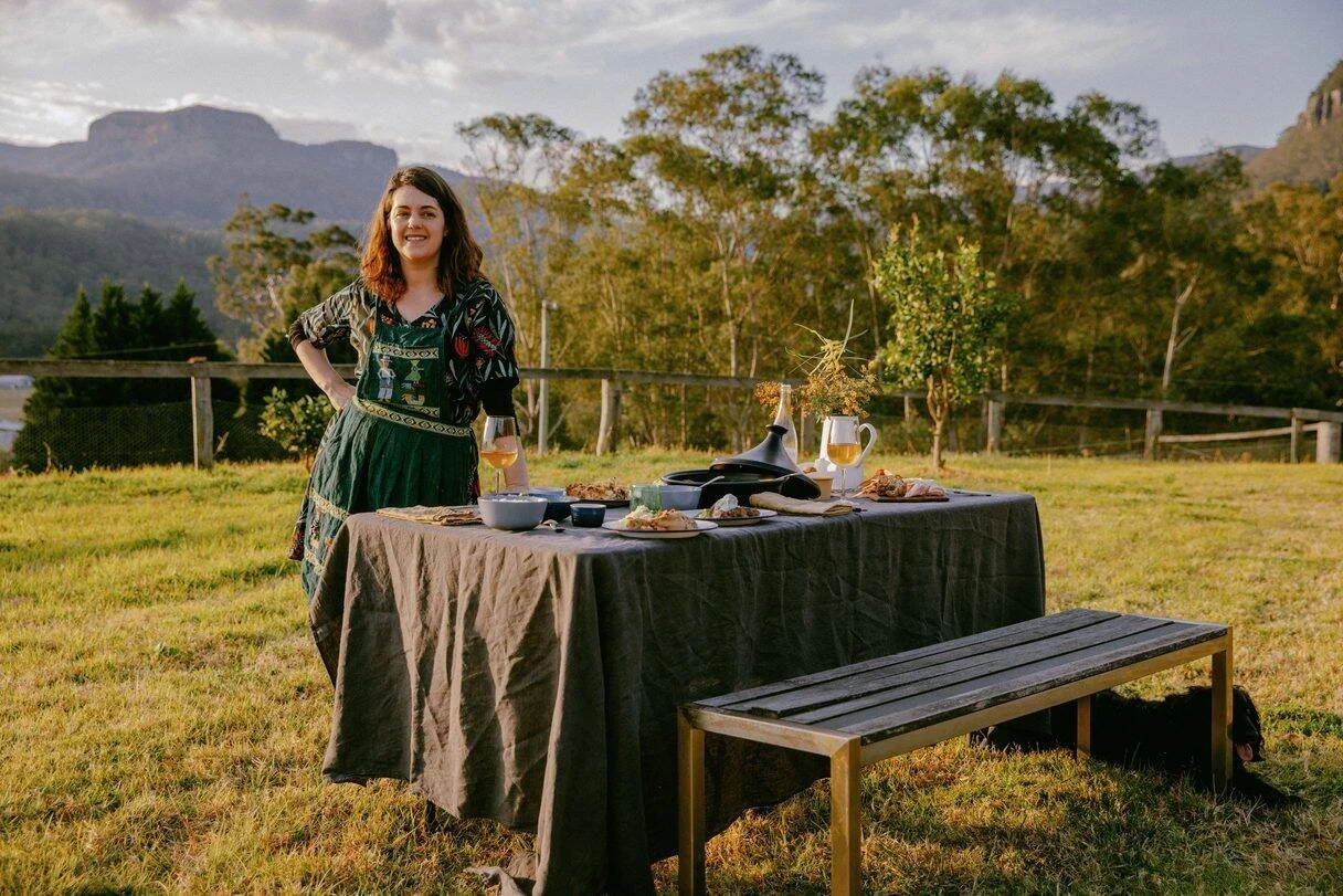 A woman standing outdoors next to a table set for a meal, with a scenic mountain and tree background during sunset.