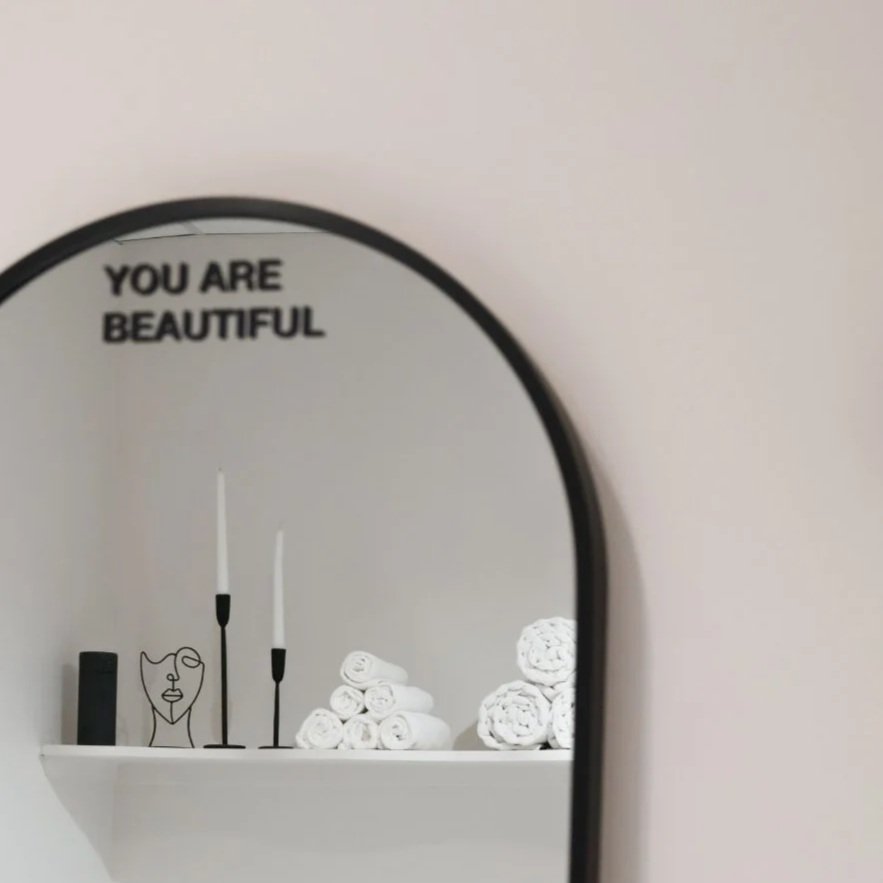 Mirror with the words "You are beautiful" on the top left corner, reflecting a shelf with rolled towels, a small black vase, a wire art face sculpture, and candles.