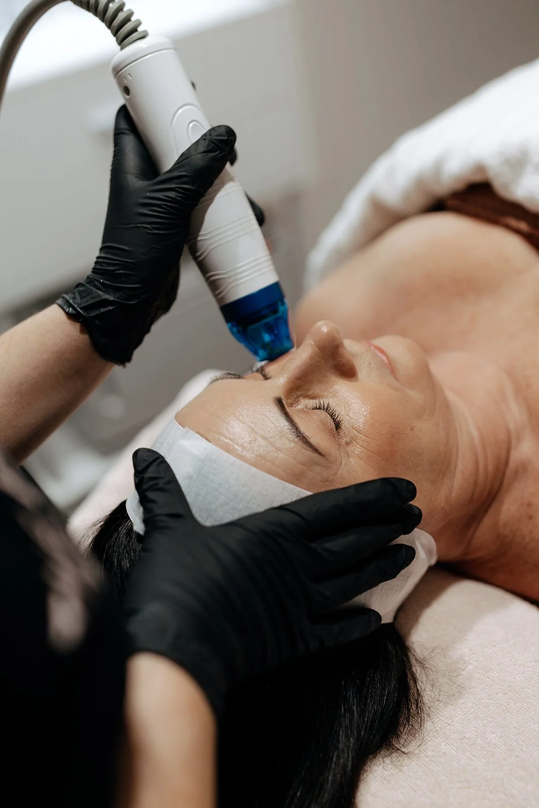 A woman receiving a facial treatment with a handheld device at a spa or clinic, lying down with a facial headband, and professional wearing black gloves administering the procedure.