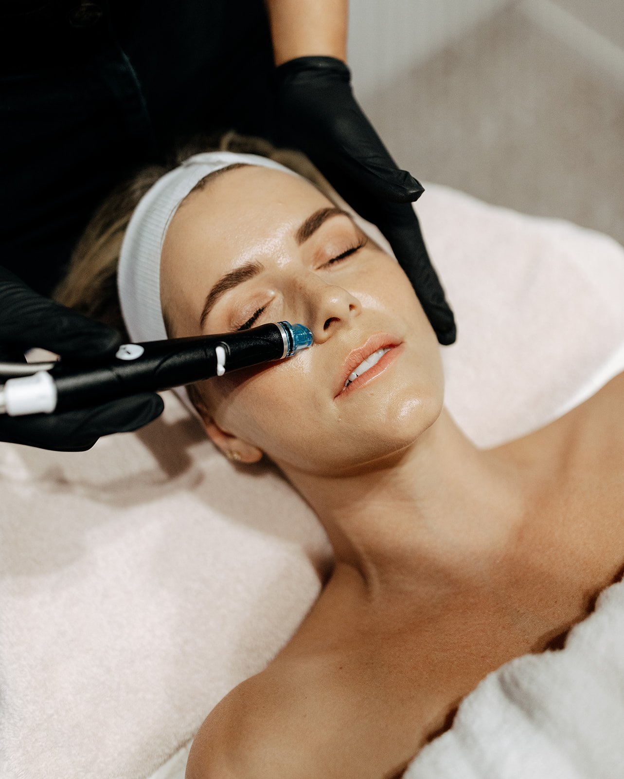 A woman receiving a facial treatment with a black pen-shaped device on her cheek, lying down with closed eyes, wearing a white headband, in a spa or clinic setting.