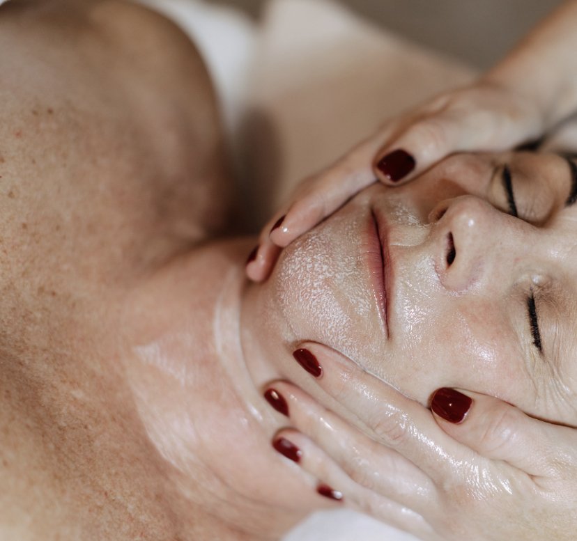 A woman receiving a spa facial massage, lying on her back with eyes closed, face covered with skincare product, and hands gently massaging her face.