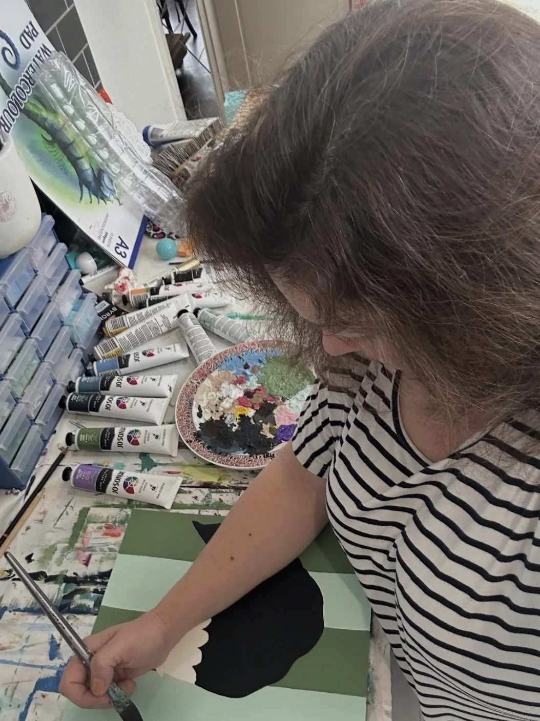 Woman painting a black silhouette on a green and light green striped background. Art supplies, including tubes of paint and a paint palette, are on the table.