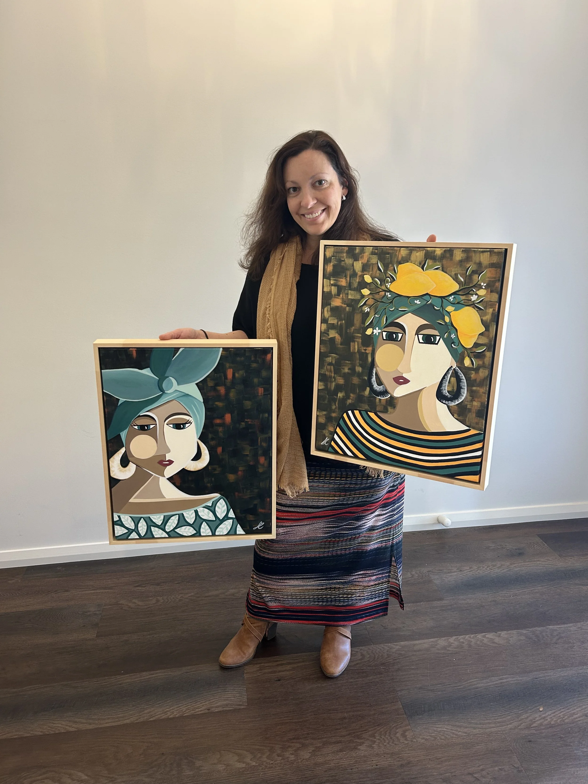 A woman with long brown hair, wearing a black top, a beige scarf, a striped skirt, and brown boots, stands indoors against a white wall, holding two framed colorful portraits of women with stylized facial features. The portraits depict women with bold features and vibrant accessories, one with a blue headscarf and the other with yellow flowers on her head.