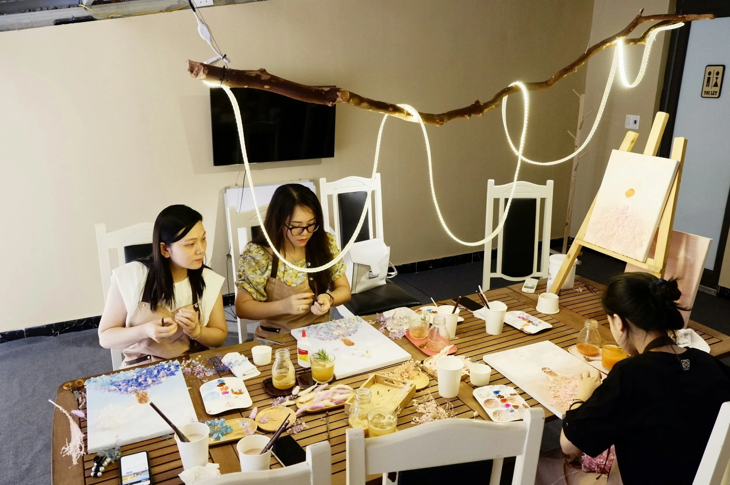 People participating in a crafting or painting workshop, seated around a table covered with art supplies such as paint palettes, brushes, and flowers, in an indoor setting with chairs and a decorative lighted branch overhead.