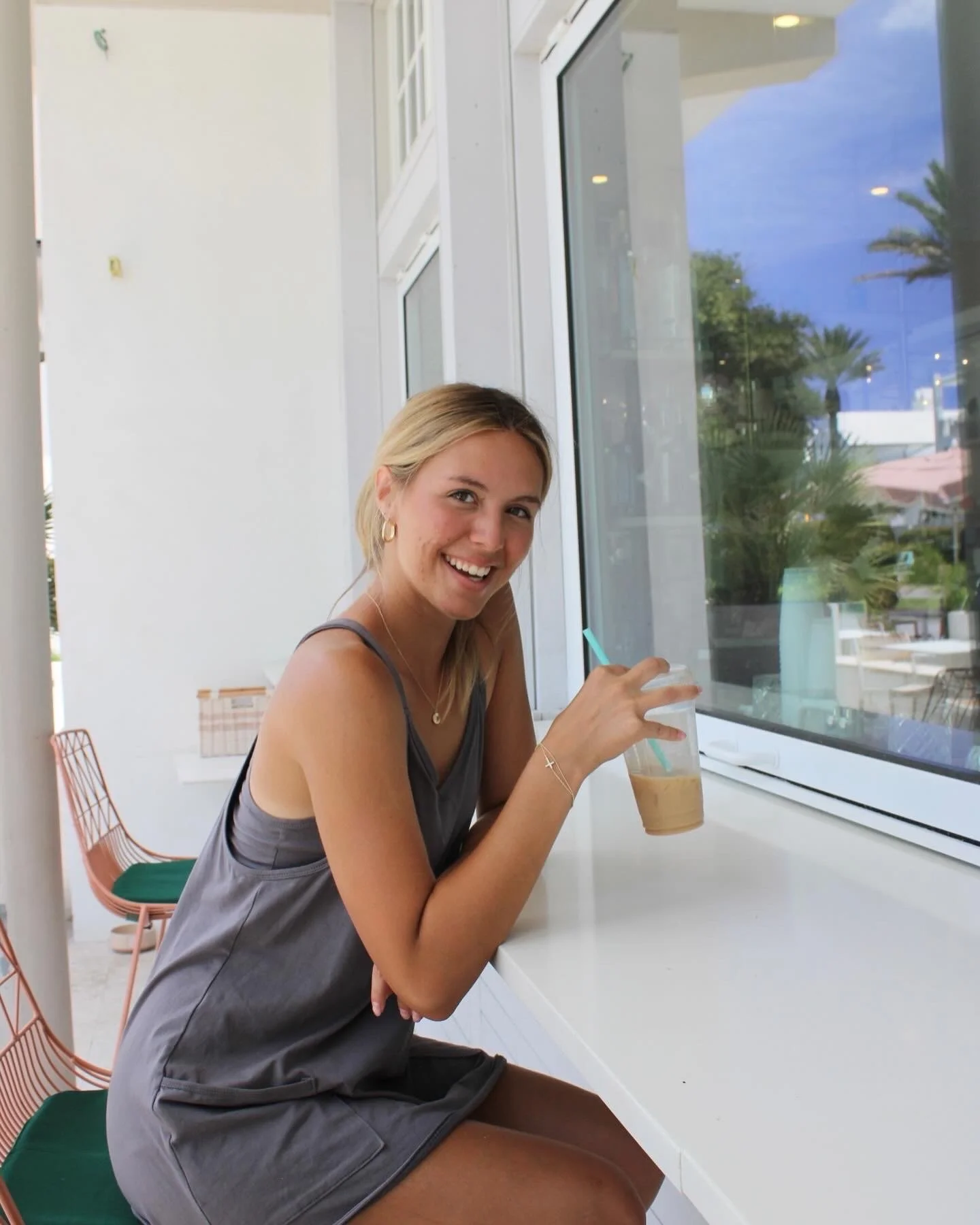 A young woman sitting at a counter enjoying a iced coffee, with a window behind her showing palm trees and a blue sky.