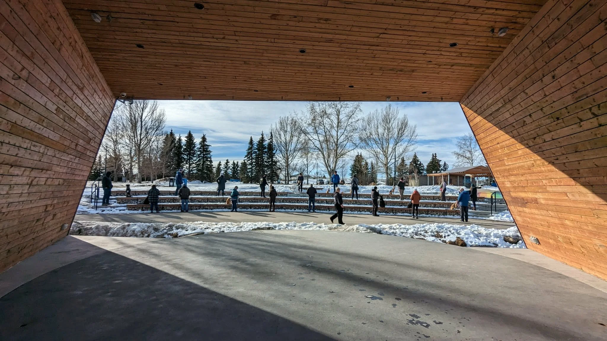 People gathered outside in a snowy park, viewed through a wooden shelter, with trees and cloudy sky in the background.