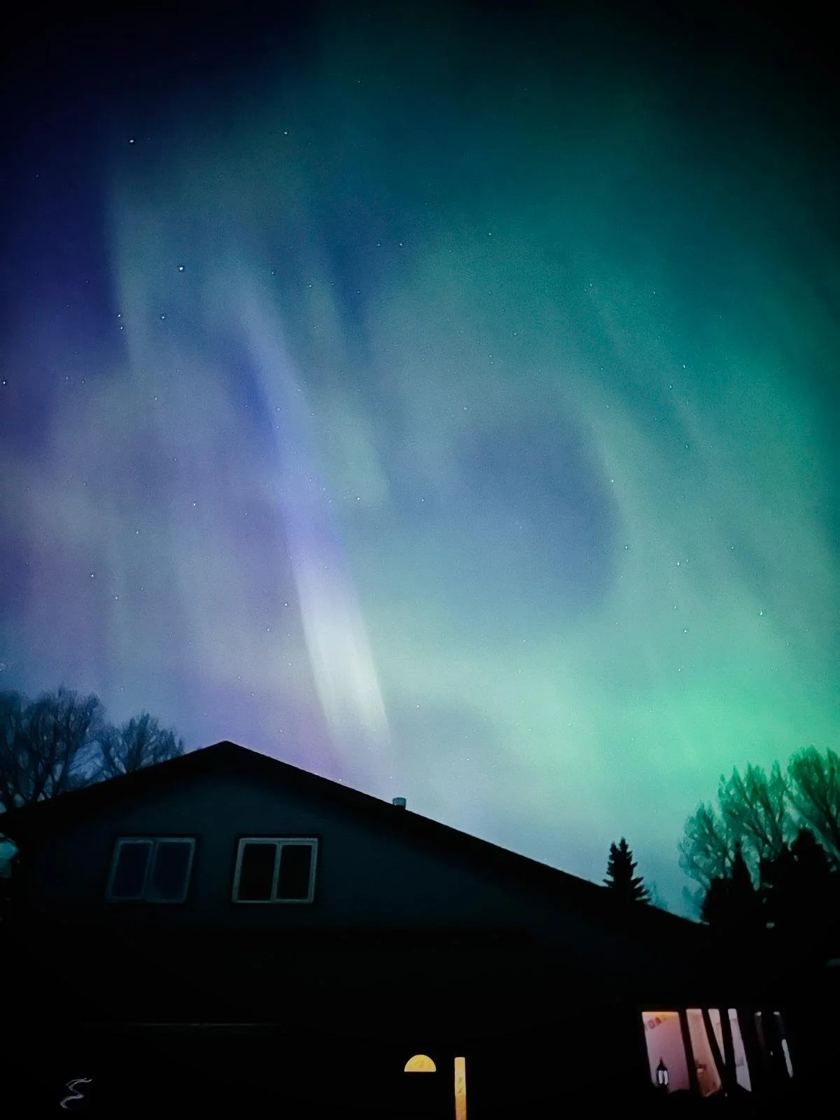 Night sky illuminated by the northern lights with visible stars, seen above a house with a dark roof and silhouette of trees in the foreground.