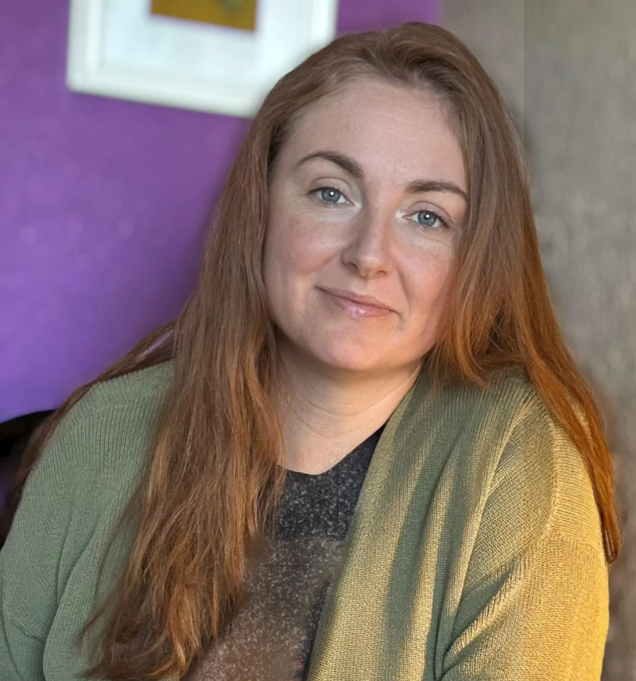 A woman with long red hair, blue eyes, and a light complexion, smiling slightly, wearing a green cardigan over a dark top, sitting indoors with a purple wall and a framed picture behind her.