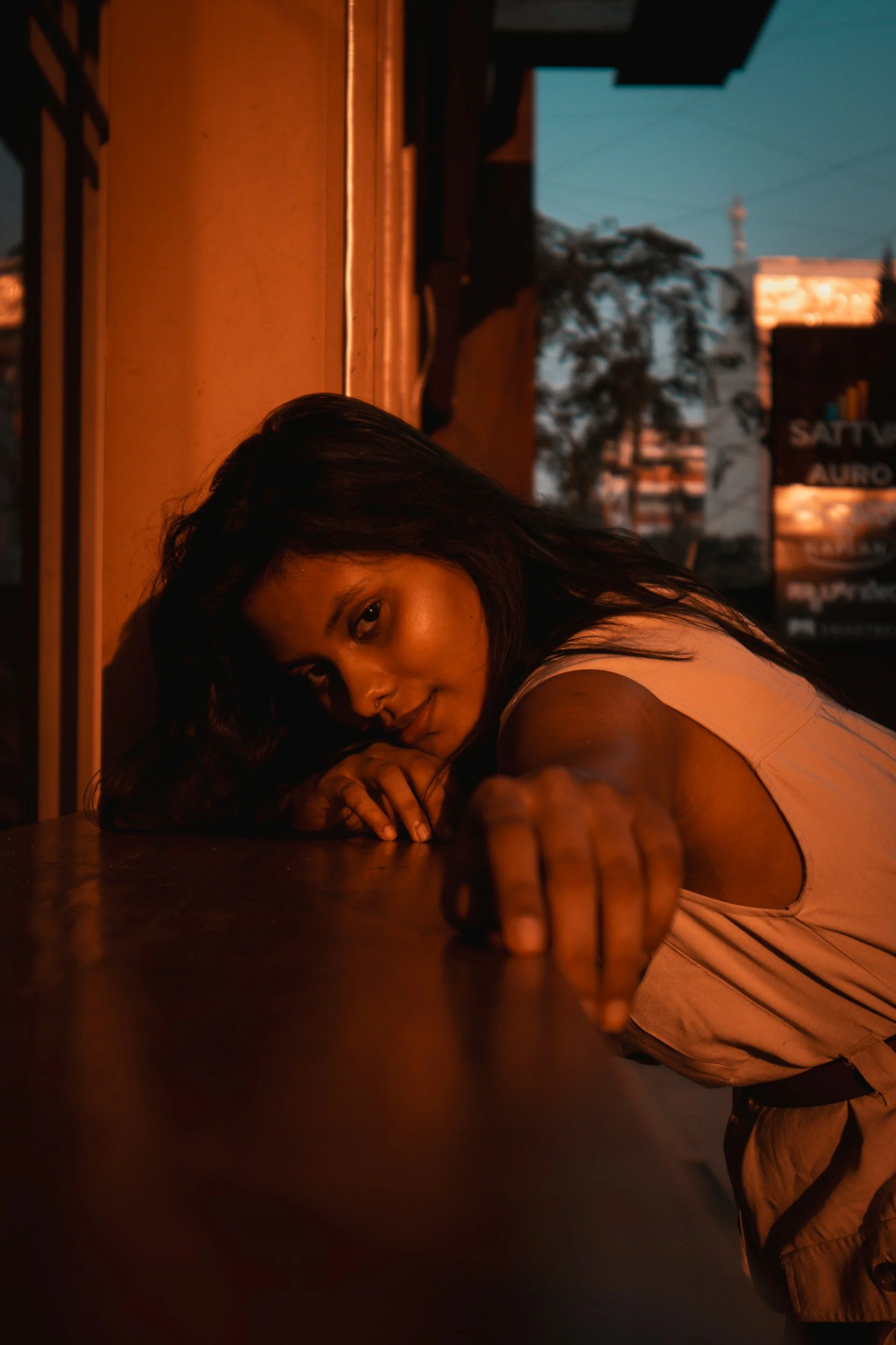 A woman with long dark hair is lying on her stomach at a table, resting her head on her crossed arms. She is in a dimly lit setting with warm lighting and an outdoor view of trees and buildings visible through the window.