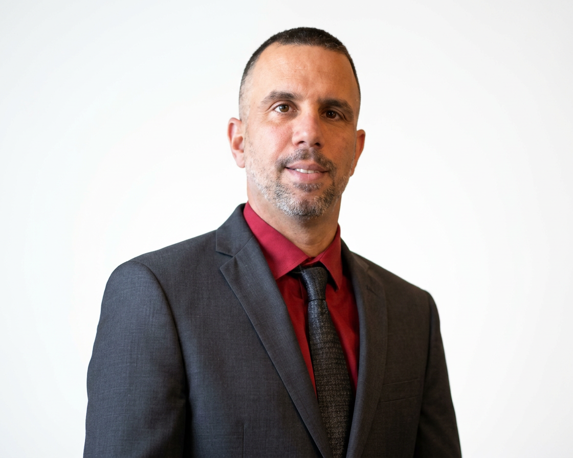 Professional man with short dark hair and beard wearing a dark suit, red shirt, and dark tie, standing against a plain white background.