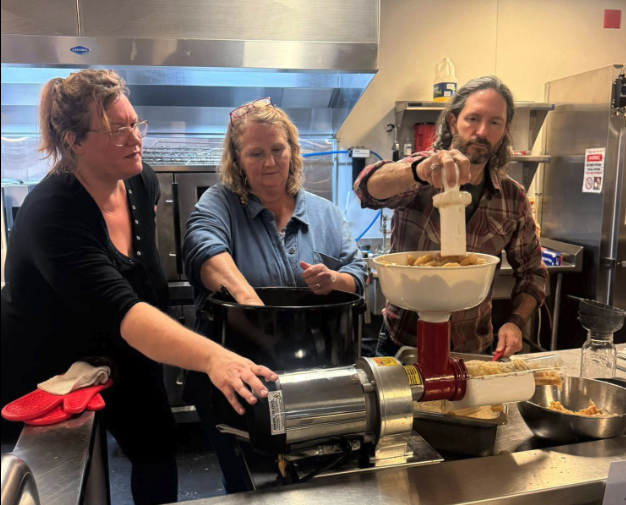 Three people in a kitchen preparing food using a manual meat grinder, with two women and one man involved in the process.