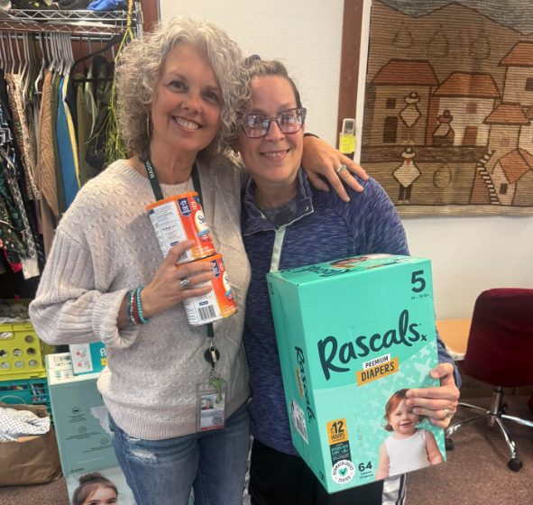Two women smiling, one holding a box of Rascals diapers, in a room with clothing and a tapestry on the wall.