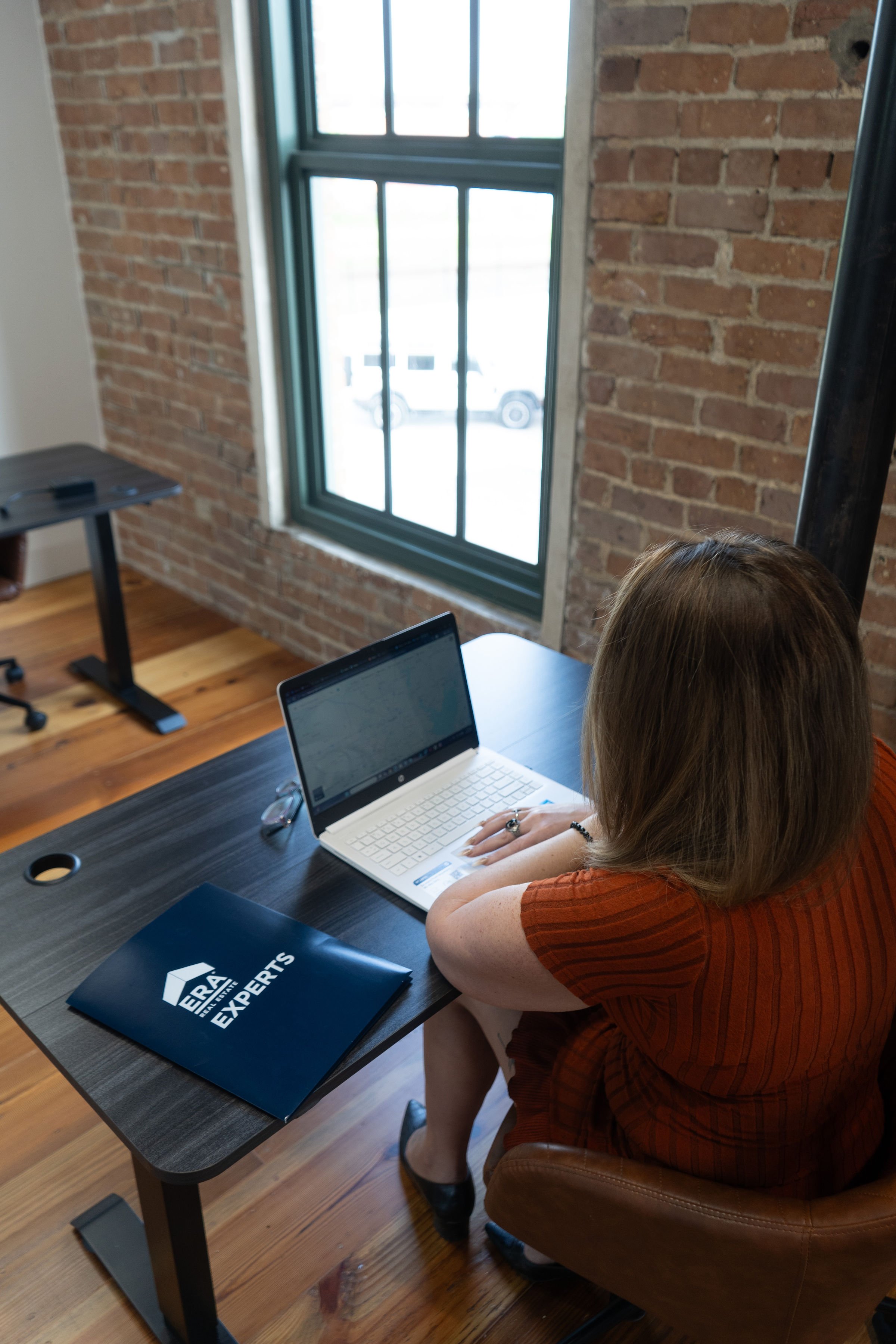 A woman sitting at a black table using a white laptop, with a booklet titled 'General Experts' on the table, in a room with brick walls and wood flooring, near a large window.