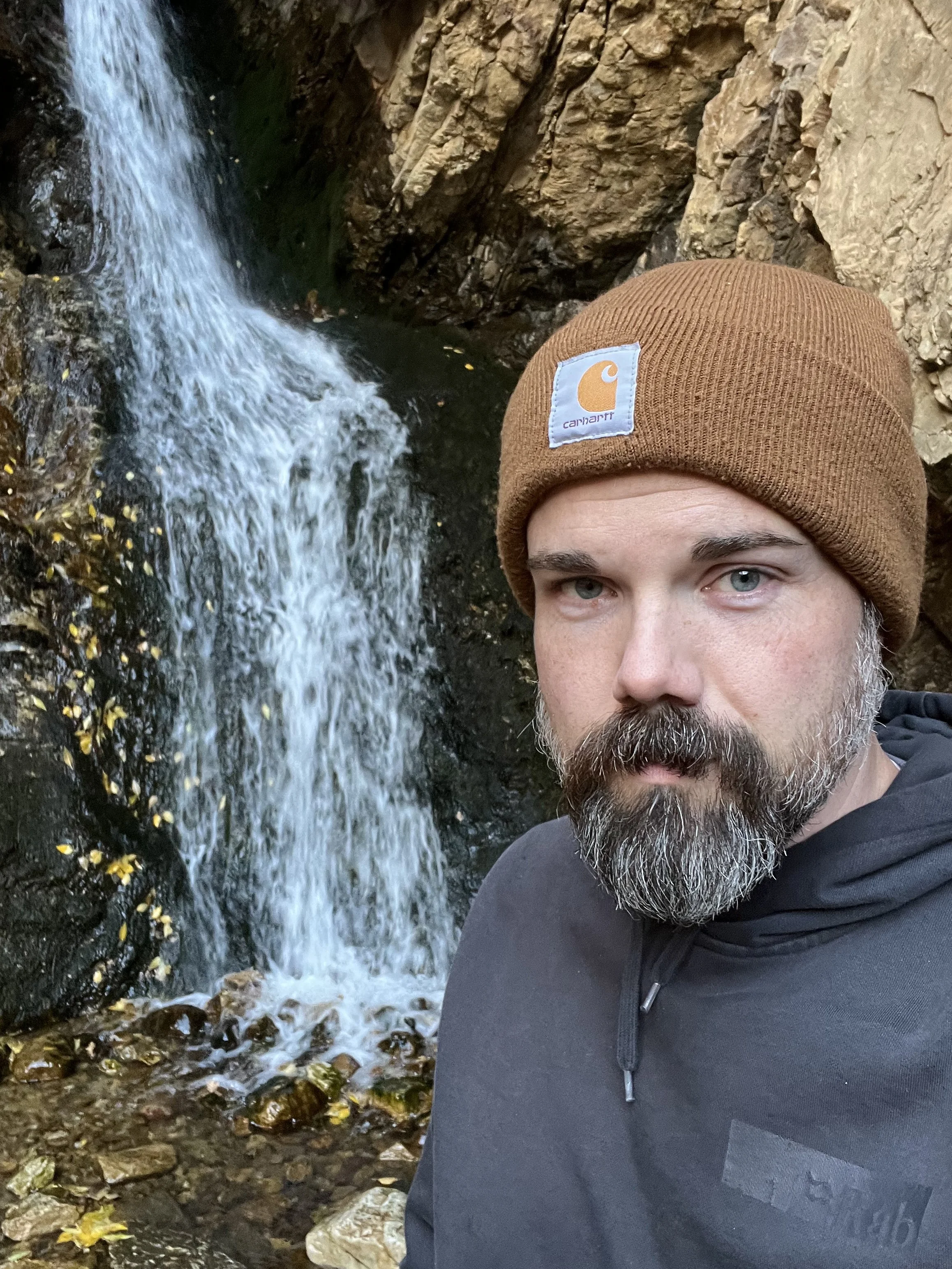 A man wearing a brown Carhartt knit hat and a black jacket taking a selfie near a small waterfall and rocky stream.