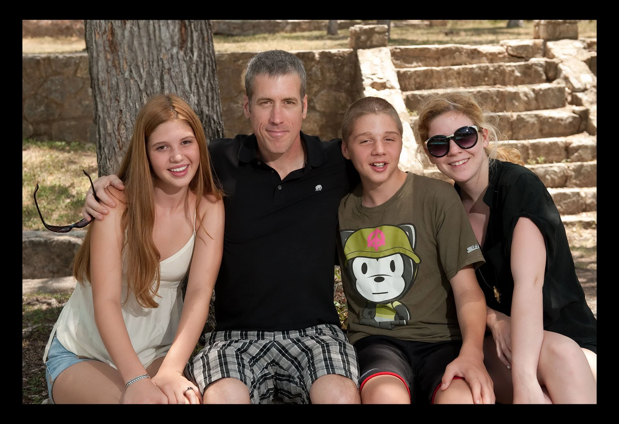 Group of four people sitting on grass outdoors, smiling, with tree and stone steps in background