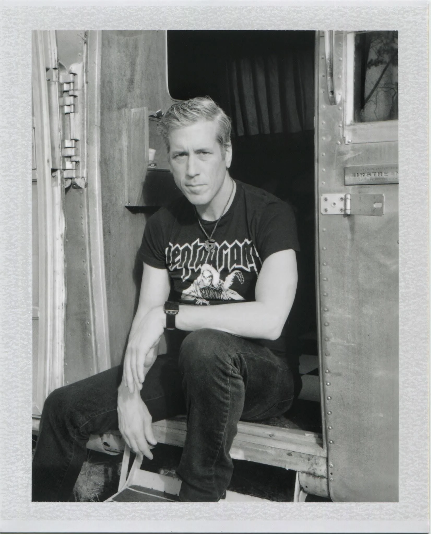 A young man with short hair and wearing a T-shirt with graphics, sitting outdoors next to a large animal, possibly a dog, with its tail resting on his lap. They are beside an old bus or trailer with an open door and visible rivets. The background has some trees and an open sky.