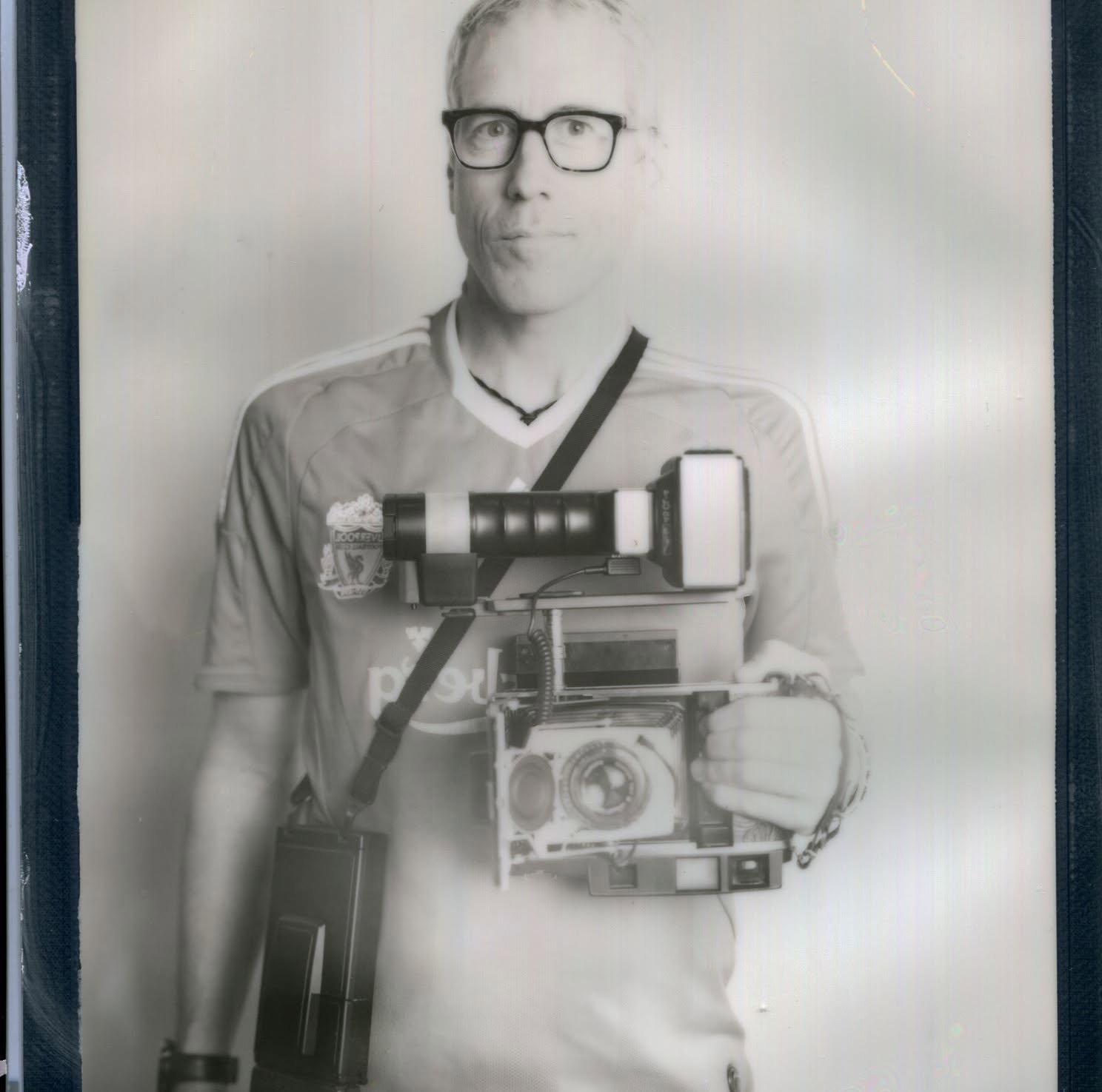 A black and white photo of a person with glasses wearing a sports jersey, holding a camera with a large stabilizer rig. The person appears to be taking a selfie.