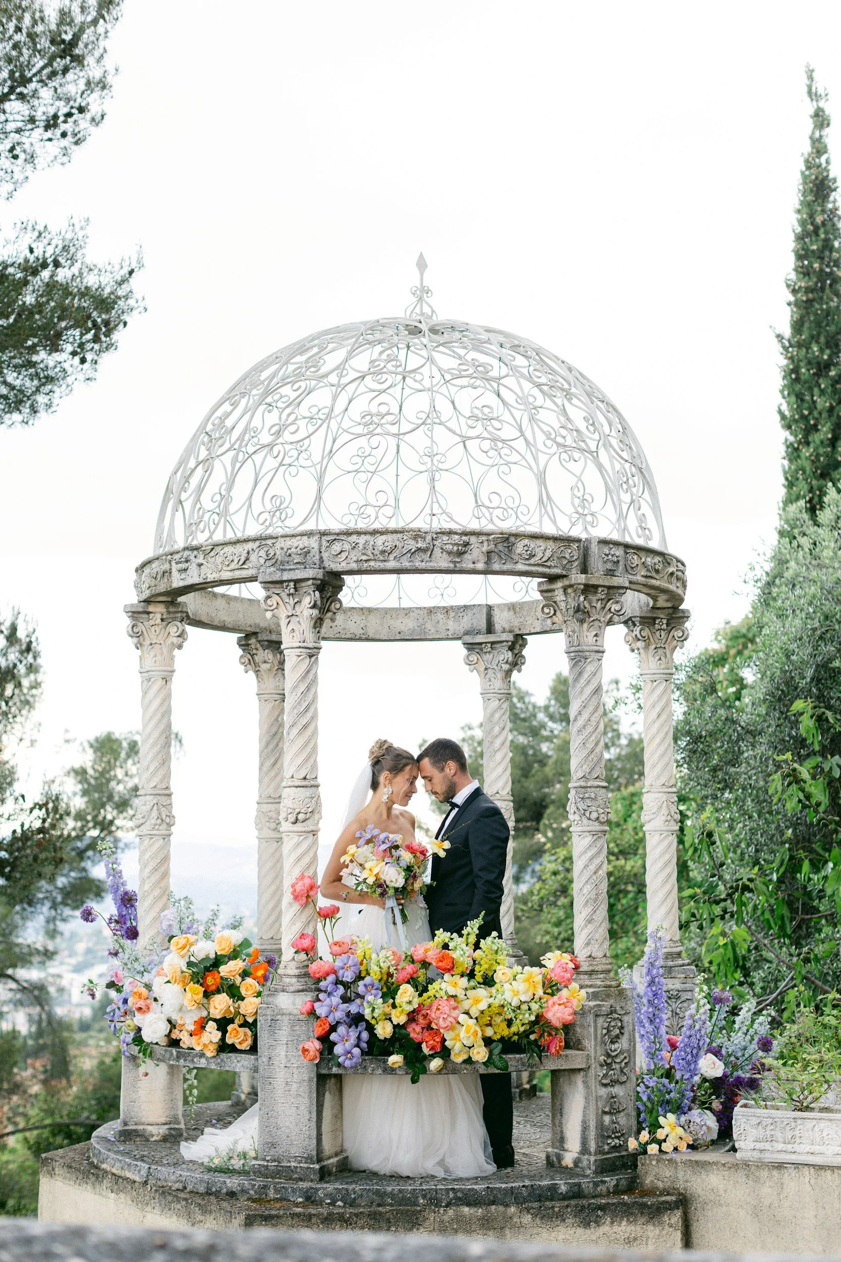 Bride and groom kissing beneath an ornate garden gazebo surrounded by colorful flowers.